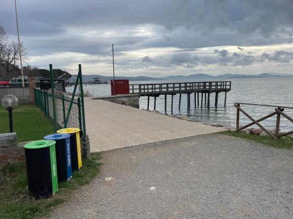 Accesso al lago con pontile in legno. A sinistra recinzione metallica verde e bidoni per rifiuti. A destra una staccionata interrotta.